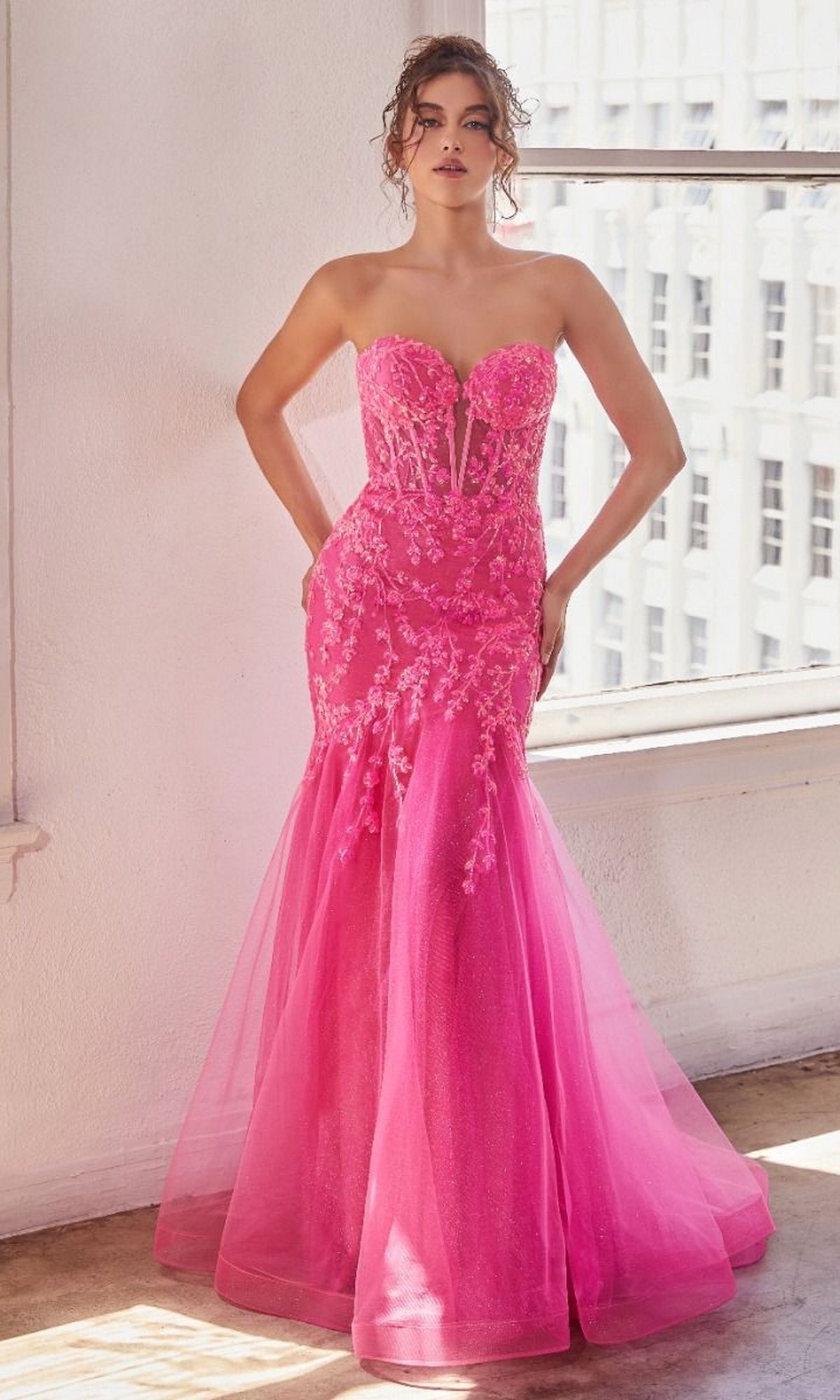 A woman stands indoors by a window wearing the Strapless Sweetheart Long Mermaid Prom Dress CB139 in bright pink, with lace and mesh details, and her hands on her hips.